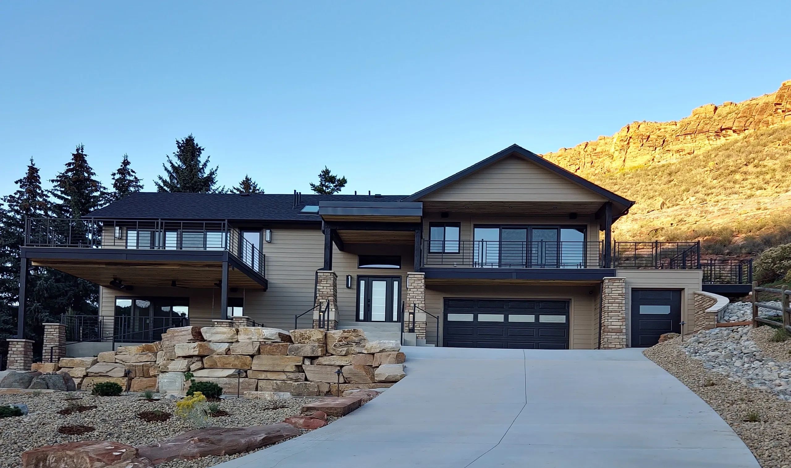 Modern two-story house with large deck, stone landscaping, and double garage at sunset